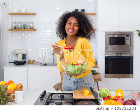 A black woman stand in a kitchen, a variety of fresh vegetable spread across the counter before her. A black woman stand in a kitchen, a variety of fresh vegetable spread across the counter before her. 108721231