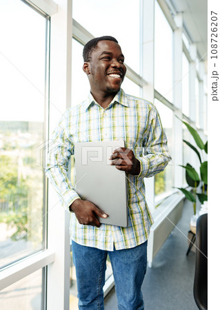 Young african man standing near the window indoors and holding laptop Young african man standing near the window indoors and holding laptop 108726207