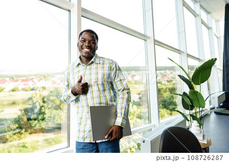 Young african man standing near the window indoors and holding laptop 108726287