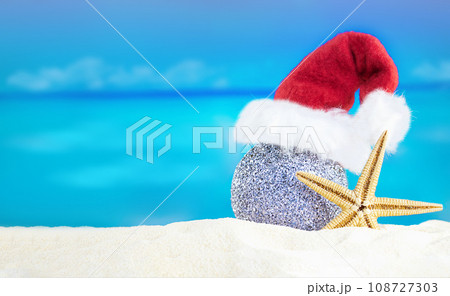 Shiny silver ball in Santa's hat with starfish on sand of beach. Christmas, New Year. Copy space Shiny silver ball in Santa's hat with starfish on sand of beach. Christmas, New Year. Copy space 108727303