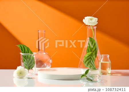 Empty white podium surrounded by laboratory glassware containing leaves and flowers. Mock-up for exhibitions or presentation of cosmetic products or packaging 108732411