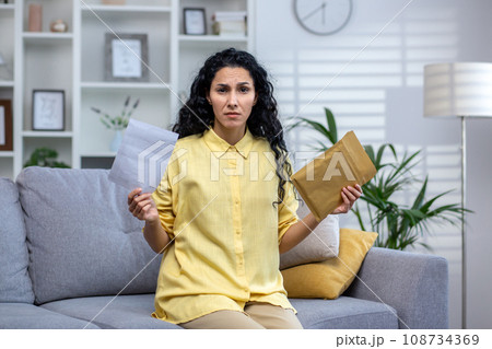 Annoyed curly haired hispanic woman sitting at home on sofa in living room holding open envelope. A cute brunette is upset about the news, losing the lottery, bad news. Annoyed curly haired hispanic woman sitting at home on sofa in living room holding open envelope. A cute brunette is upset about the news, losing the lottery, bad news. 108734369