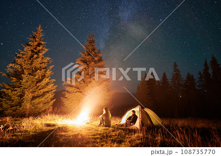 Night camping in mountains under starry sky. Two people, couple having a rest near burning campfire, enjoying beautiful sky full of stars. Concept of tourism and healthy living. 108735770