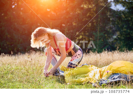 Woman hiker camping outdoors. Young female tourist putting tent, wearing sportswear, traveling in mountains. Concept of tourism and beauty of nature. 108735774
