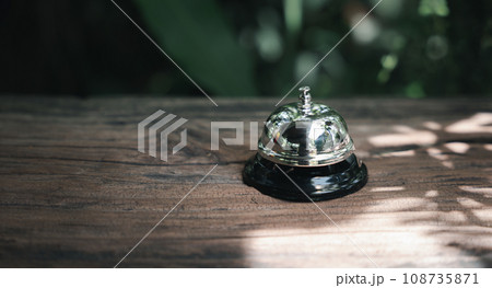 Hotel ring bell. Closeup of silver service restaurant bell on wooden counter desk, vintage bell to call staff outdoor in garden with green leaf Hotel ring bell. Closeup of silver service restaurant bell on wooden counter desk, vintage bell to call staff outdoor in garden with green leaf 108735871