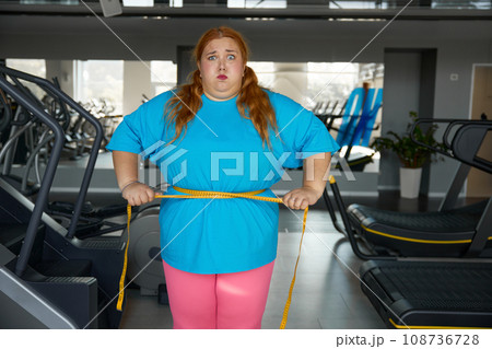 Portrait cute funny woman with excess weight ties herself with measuring tape Portrait cute funny woman with excess weight ties herself with measuring tape 108736728