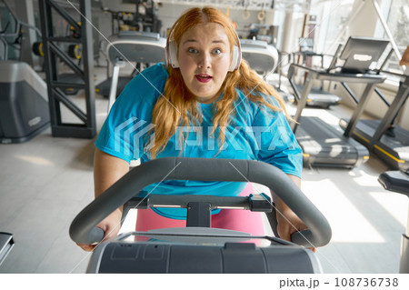 Portrait of motivated overweight woman riding exercise bike at gym 108736738