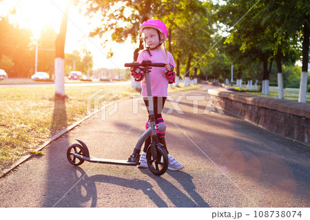 a beautiful girl in a pink helmet and protective equipment stands with a scooter on an alley in the city against the backdrop of a sunny sunset in summer. 108738074
