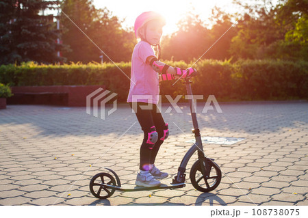 A beautiful caucasian girl of seven years old rides a scooter in a pink outfit, a helmet and knee pads on a sunny summer day. Safety in active sports. A beautiful caucasian girl of seven years old rides a scooter in a pink outfit, a helmet and knee pads on a sunny summer day. Safety in active sports. 108738075