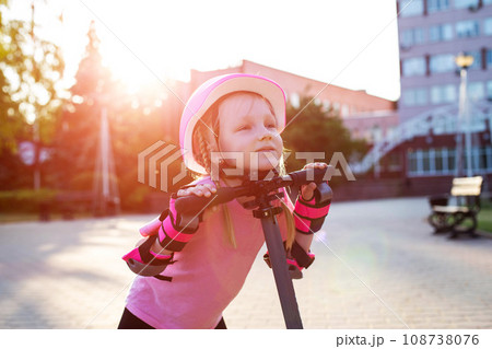 A beautiful caucasian girl of seven years old in a pink protective helmet and outfit is fooling around on a scooter in the summer. Smiling girl laughs next to the scooter. Sunset A beautiful caucasian girl of seven years old in a pink protective helmet and outfit is fooling around on a scooter in the summer. Smiling girl laughs next to the scooter. Sunset 108738076