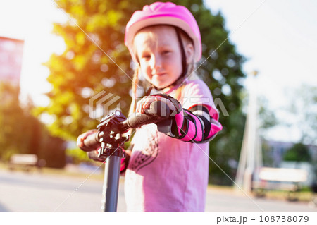 A beautiful caucasian girl of 7 years old in a pink helmet and armlets stands on a scooter. Summer sunny day. 108738079