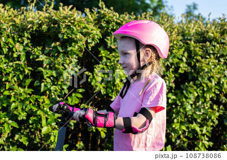 A beautiful girl of 7 years old in a pink helmet and protective sleeves stands against the background of green bushes with a scooter. Safe scooter riding. A beautiful girl of 7 years old in a pink helmet and protective sleeves stands against the background of green bushes with a scooter. Safe scooter riding. 108738086