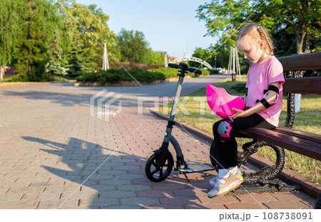 A 7-year-old girl sits on a bench in protective knee pads and holds a helmet in her hands. Scooter injury protection kit. Copy space for text A 7-year-old girl sits on a bench in protective knee pads and holds a helmet in her hands. Scooter injury protection kit. Copy space for text 108738091