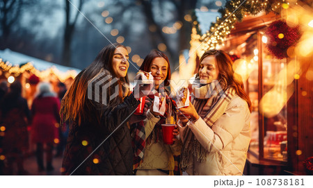 Happy young women attending winter fair in evening, drinking hot drinks, eating hot dogs. Merry season and cheerful time Happy young women attending winter fair in evening, drinking hot drinks, eating hot dogs. Merry season and cheerful time 108738181