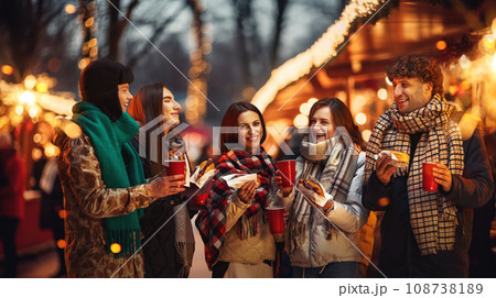 Happy, laughing people, friends standing on outdoor winter fair, drinking hot drinks, eating hot dogs. Merry season 108738189