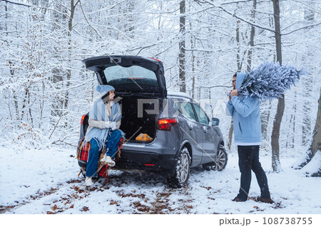In a winter forest, a woman sits in the trunk of a car, and a man carries a Christmas tree In a winter forest, a woman sits in the trunk of a car, and a man carries a Christmas tree 108738755