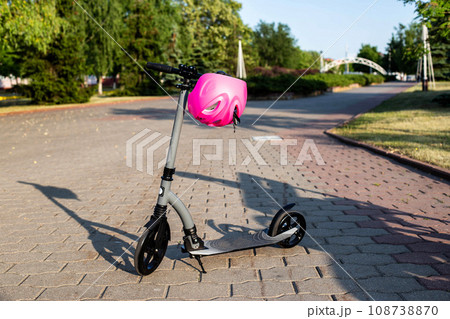 A modern scooter stands on an alley with a pink helmet against the backdrop of a sunset in summer. The concept of safety while riding a scooter. Copy space for text A modern scooter stands on an alley with a pink helmet against the backdrop of a sunset in summer. The concept of safety while riding a scooter. Copy space for text 108738870