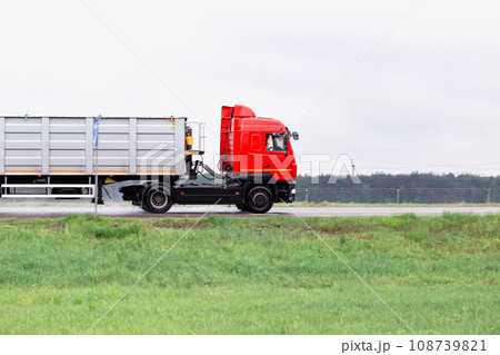 A red truck with a semi-trailer transports bulk cargo along the road in cloudy weather. Side view. Copy space for text, industry A red truck with a semi-trailer transports bulk cargo along the road in cloudy weather. Side view. Copy space for text, industry 108739821