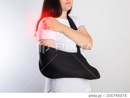 A girl on a white background with a black supporting medical bandage after a dislocation of the shoulder joint and a bone fracture. Rehabilitation after injury, orthopedics and traumatology 108740078