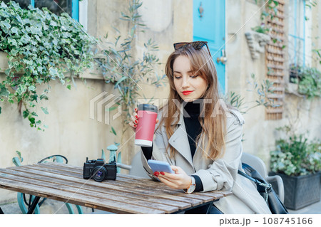 Female traveler chatting, sharing files, resting in cafe terrace table with vintage camera and coffee in reusable cup after city walk. Girl using internet in wifi zone, enjoying coffee and leisure 108745166