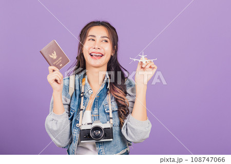 Happy Asian portrait beautiful young woman excited smiling in summer vacation holding airplane toy, passport and vintage photo camera, Thai female ready travel trip isolated on purple background 108747066