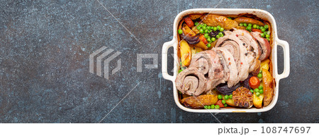 Rolled sliced pork roasted in white casserole dish with potatoes, vegetables and herbs on blue dark concrete rustic background top view. Baked pork roll with vegetables for dinner 108747697