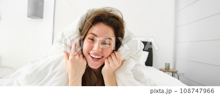 Portrait of beautiful brunette woman lying in bed, resting in her hotel room, covered in white blankets, laughing and smiling at camera Portrait of beautiful brunette woman lying in bed, resting in her hotel room, covered in white blankets, laughing and smiling at camera 108747966