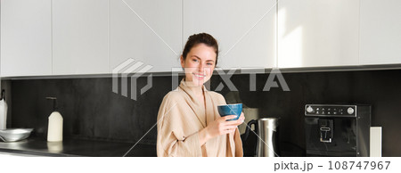 Daily routine and lifestyle. Young beautiful woman in bathrobe, standing in kitchen with cup of coffee, drinking tea, smiling and looking happy 108747967