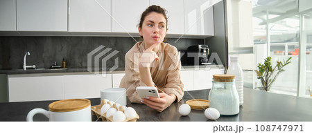 Attractive young cheerful girl baking at the kitchen, making dough, holding recipe book, having ideas Attractive young cheerful girl baking at the kitchen, making dough, holding recipe book, having ideas 108747971
