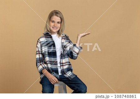 Cute blonde teen girl in checkered shirt sitting on the chair in a studio 108748861