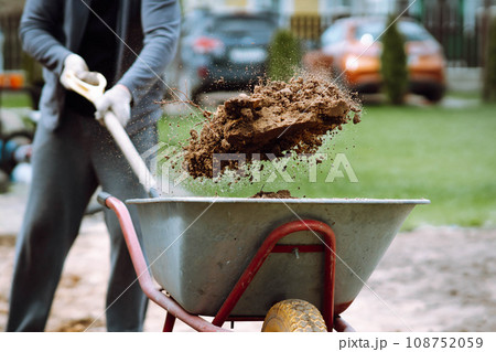 Gardener in gloves throw collected earth with spade into wheelbarrow. Man in garden dig ground in morning. Gardener in gloves throw collected earth with spade into wheelbarrow. Man in garden dig ground in morning. 108752059