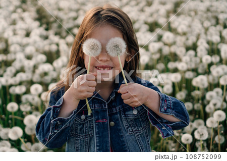 Joyful girl standing in field of white dandelions and holding two flowers in hands covering eyes with fluffy blowballs. Joyful girl standing in field of white dandelions and holding two flowers in hands covering eyes with fluffy blowballs. 108752099