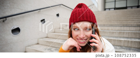 Beautiful smiling redhead female model, sits on street and talks on mobile phone, uses smartphone app to call abroad, laughing during telephone conversation 108752335