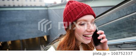 Portrait of young redhead woman walks around city, goes up stairs with mobile phone, talks on smartphone and smiles Portrait of young redhead woman walks around city, goes up stairs with mobile phone, talks on smartphone and smiles 108752337