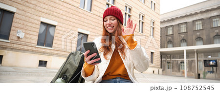 Social media and people. Young redhead girl sits on street, uses mobile phone app, looks up information in internet, holds smartphone Social media and people. Young redhead girl sits on street, uses mobile phone app, looks up information in internet, holds smartphone 108752545