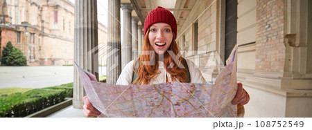 Adventurous redhead girl walks in town with paper map, explores city as tourist, looks for popular tourism attractions, looks around excited and smiles 108752549
