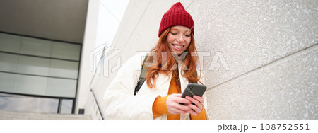 Happy girl student in red hat, holds smartphone, tourist looks at map app on her phone, explores sightseeing, texts message, looks for couchsurfing, rents place to stay online Happy girl student in red hat, holds smartphone, tourist looks at map app on her phone, explores sightseeing, texts message, looks for couchsurfing, rents place to stay online 108752551