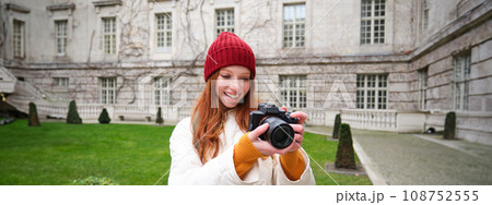 Happy redhead girl tourist, takes photos, photographer with professional camera walks around city and captures beautiful pictures Happy redhead girl tourist, takes photos, photographer with professional camera walks around city and captures beautiful pictures 108752555