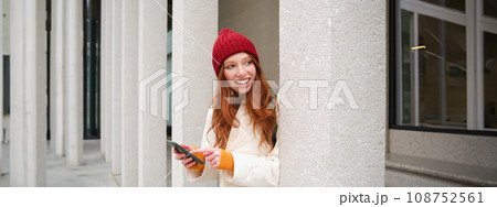 Stylish urban girl using mobile phone app, standing in city, waiting for taxi, looking at smartphone application, texting message 108752561