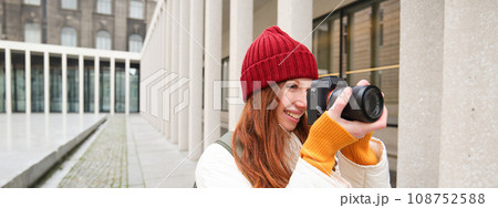 Smiling redhead girl photographer, taking pictures in city, makes photos outdoors on professional camera Smiling redhead girl photographer, taking pictures in city, makes photos outdoors on professional camera 108752588
