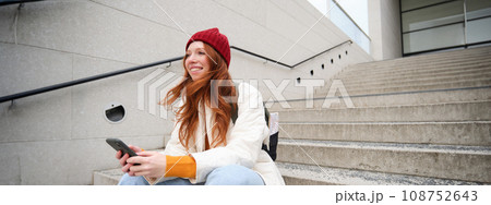 Mobile phones and people. Young stylish redhead girl sits on stairs with telephone, uses smartphone app, reads smth online Mobile phones and people. Young stylish redhead girl sits on stairs with telephone, uses smartphone app, reads smth online 108752643