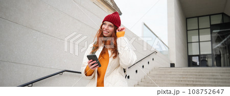 Happy girl student in red hat, holds smartphone, tourist looks at map app on her phone, explores sightseeing, texts message, looks for couchsurfing, rents place to stay online 108752647