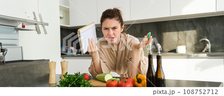 Portrait of woman cant cook, looking confused while making meal, holding recipe book, checking grocery list and staring frustrated at camera, standing near vegetables in the kitchen Portrait of woman cant cook, looking confused while making meal, holding recipe book, checking grocery list and staring frustrated at camera, standing near vegetables in the kitchen 108752712