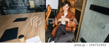 Image of young professional entrepreneur, woman in an office, sitting in chair in an office, using mobile phone, messaging a co-worker 108752802