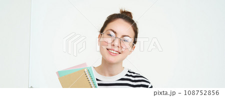 Portrait of young woman, tutor in glasses, holding her notebooks and documents, smiling at camera, standing over white background 108752856