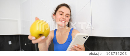 Portrait of smiling, happy fitness girl, sitting with an apple, laughing over smth on smartphone, browsing social media on mobile phone app 108753208