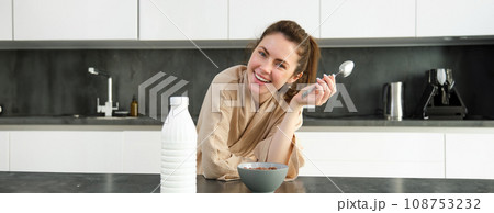 Portrait of young beautiful woman in bathrobe, eating cereals for breakfast, leans on kitchen worktop, looking at her morning meal 108753232