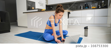 Image of young fitness instructor using laptop for online workout training, showing fitness exercises, talking to client, sitting on yoga rubber mat in sportswear Image of young fitness instructor using laptop for online workout training, showing fitness exercises, talking to client, sitting on yoga rubber mat in sportswear 108753300
