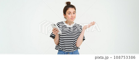 Image of clueless girl shrugs shoulders with smartphone in hand, isolated over white background Image of clueless girl shrugs shoulders with smartphone in hand, isolated over white background 108753386