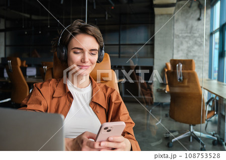 Portrait of female employee, entrepreneur in office with laptop, mobile phone, wears wireless headphones 108753528
